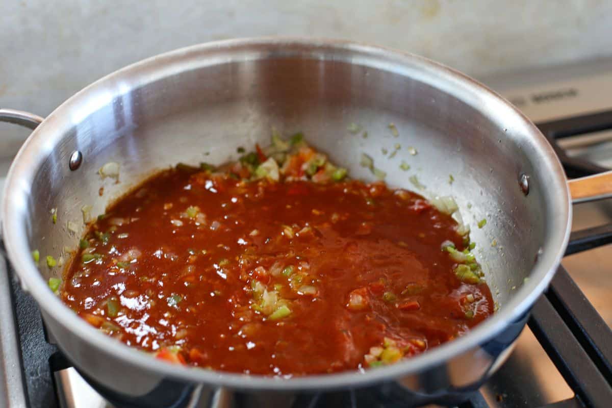 A stainless steel pan on a stove contains enchilada sauce with chopped vegetables simmering.