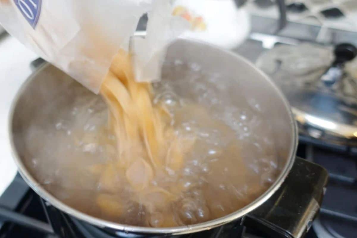 Dry pasta is being poured from a bag into a pot of boiling water on a stovetop.