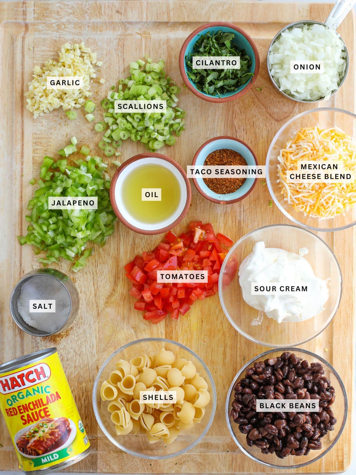 A wooden board displays labeled ingredients for a dish: garlic, scallions, cilantro, onion, jalapeno, oil, taco seasoning, cheese blend, salt, tomatoes, sour cream, shells, black beans, and enchilada sauce.