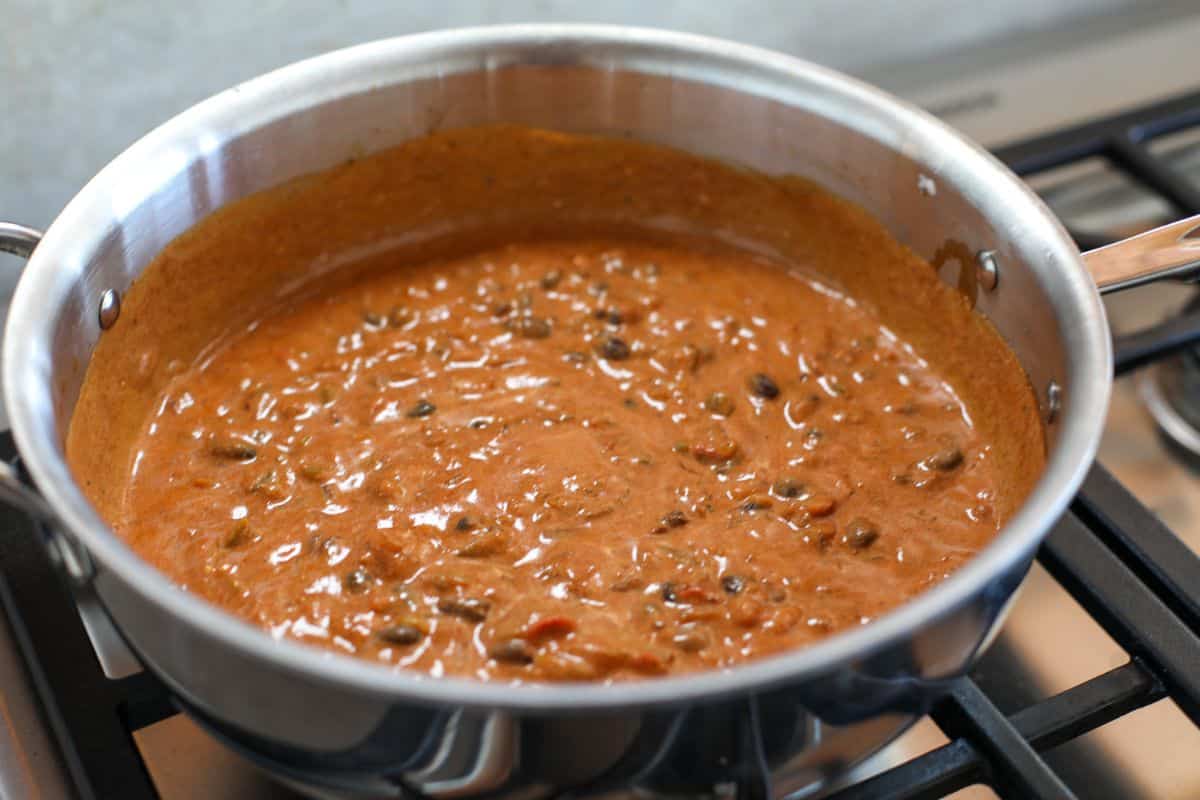 A stainless steel pan on a stove contains a thick, orange-brown enchilada sauce with visible bits of beans and spices.