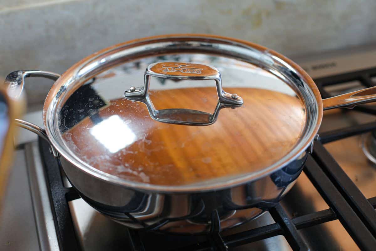 A stainless steel pan with a lid sits on a gas stove burner in a kitchen.