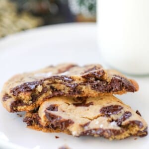 Two chocolate chip cookies broken in half are stacked on a white plate, with a glass of milk in the background.