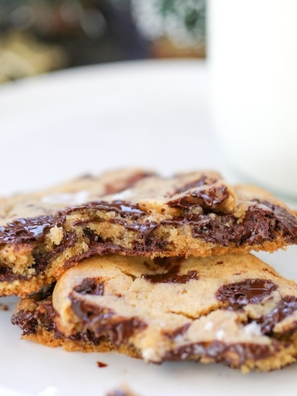 Two chocolate chip cookies broken in half are stacked on a white plate, with a glass of milk in the background.