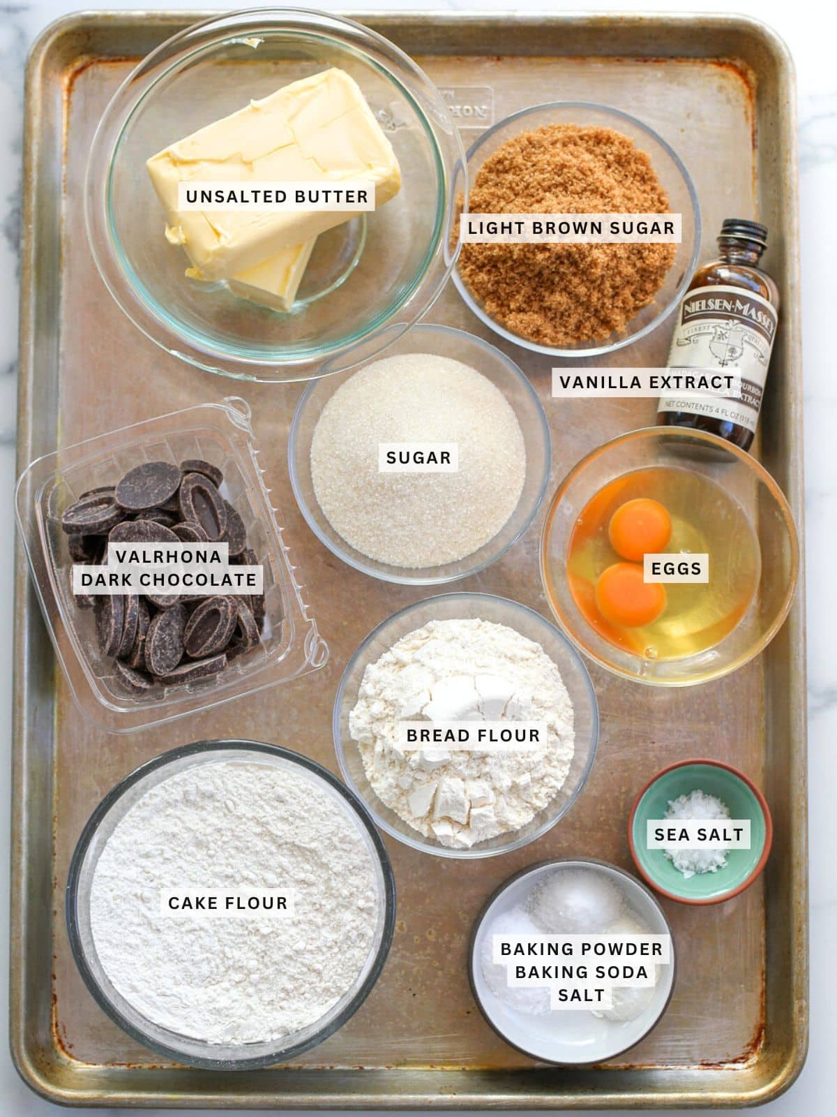 A baking sheet holds bowls of unsalted butter, light brown sugar, vanilla extract, white sugar, eggs, Valrhona dark chocolate, bread flour, cake flour, sea salt, baking powder, and baking soda.