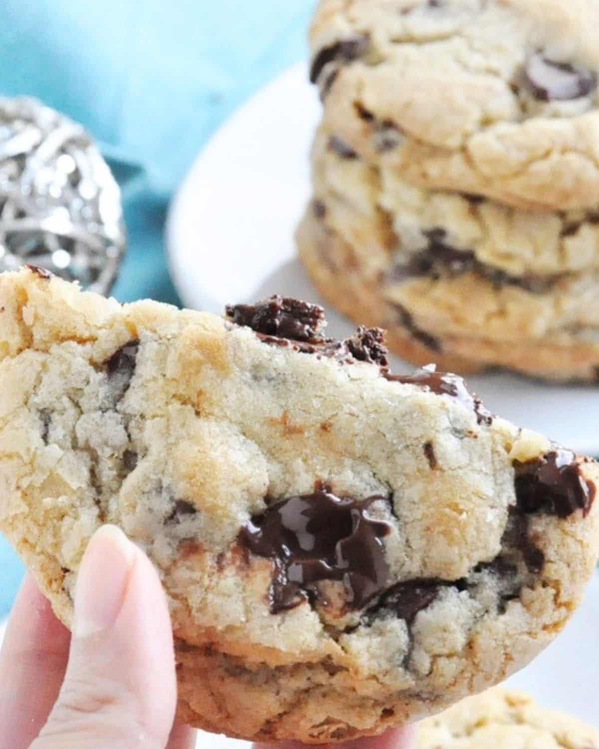A hand holding a large chocolate chip cookie with melted chocolate chunks, with more cookies stacked on a plate in the background.