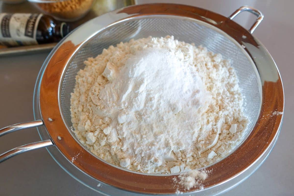 A metal sifter containing a mound of flour and baking powder, baking soda, and salt sits on a kitchen counter with vanilla extract and other ingredients in the background.
