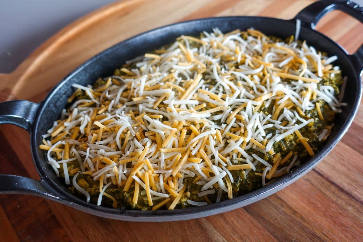 A black oval baking dish containing palak paneer topped with shredded yellow and white cheese, placed on a wooden surface.