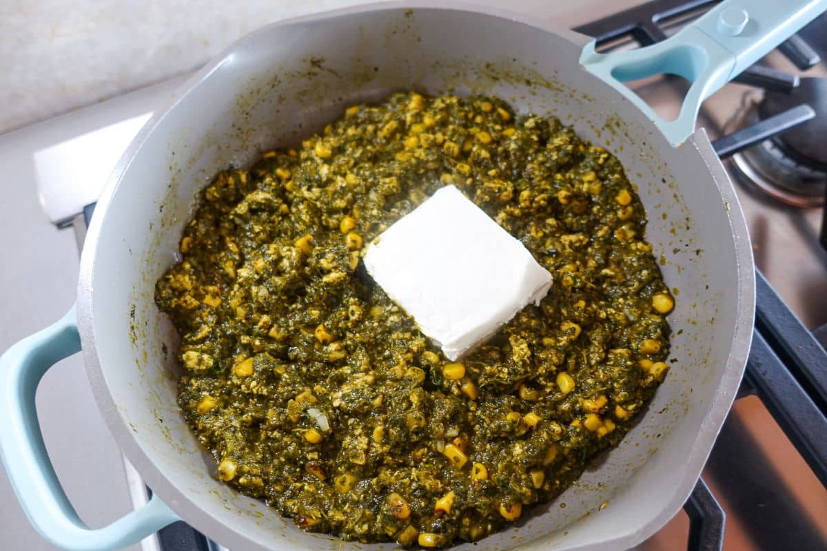 A pan of cooked palak paneer corn sabzi topped with a square block of cream cheese on a stovetop.