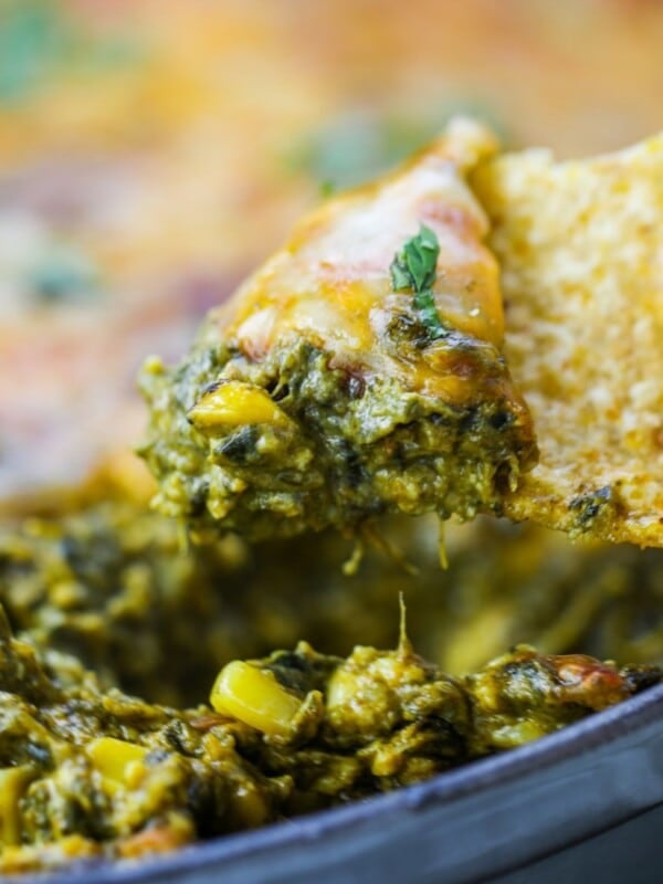 A tortilla chip scooping up cheesy palak paneer dip with corn from a baking dish.