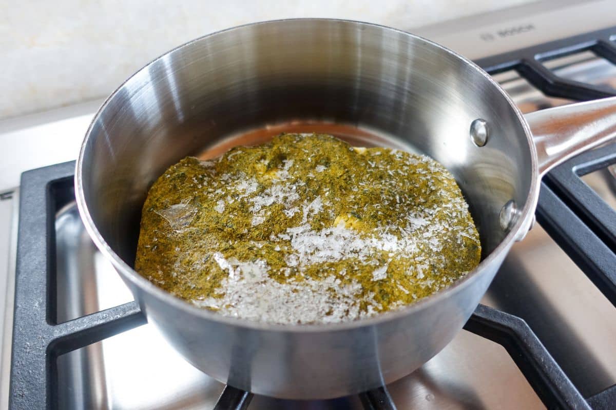 A metal saucepan containing a frozen palak paneer block with some frost on top sits on a stovetop burner.