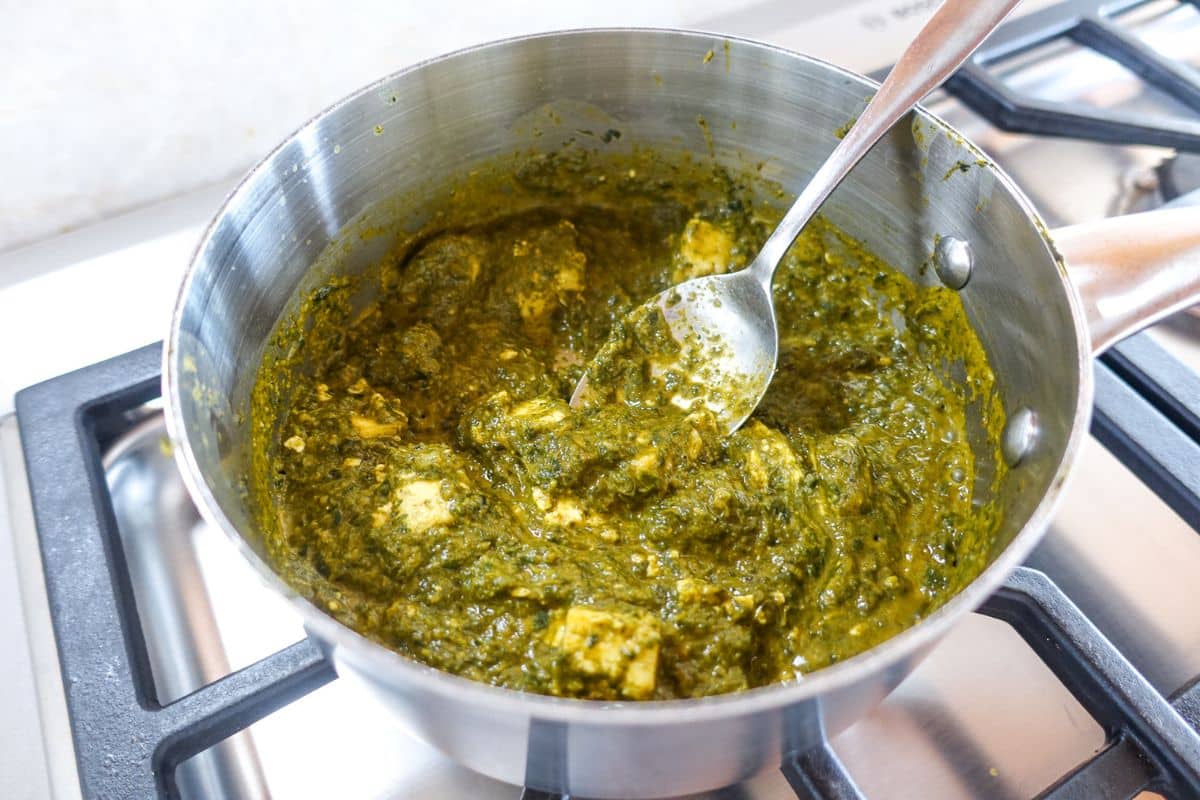 A saucepan filled with palak paneer, an Indian spinach and cheese dish, sits on a stove with a spoon inside.