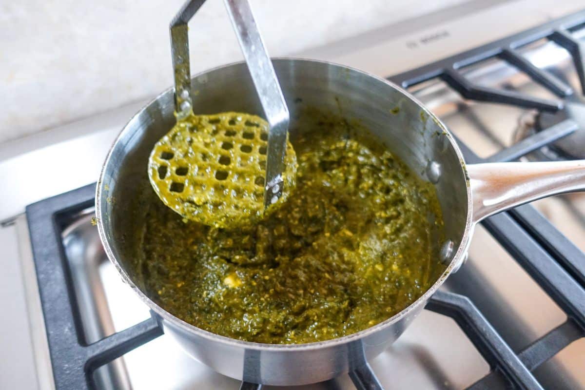 A metal pot on a stove contains palak paneer, mashed vegetable mixture being stirred with a potato masher.