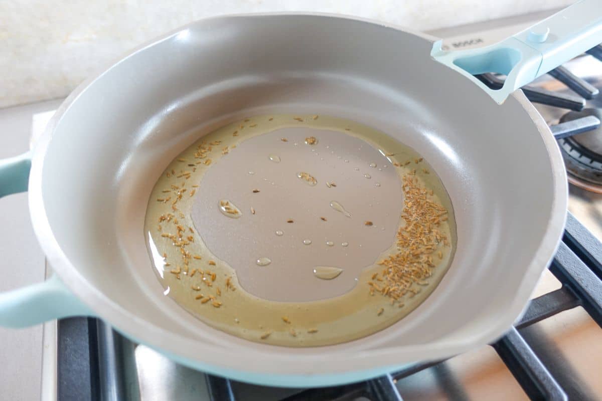 A frying pan with oil and cumin seeds heating on a stove burner.