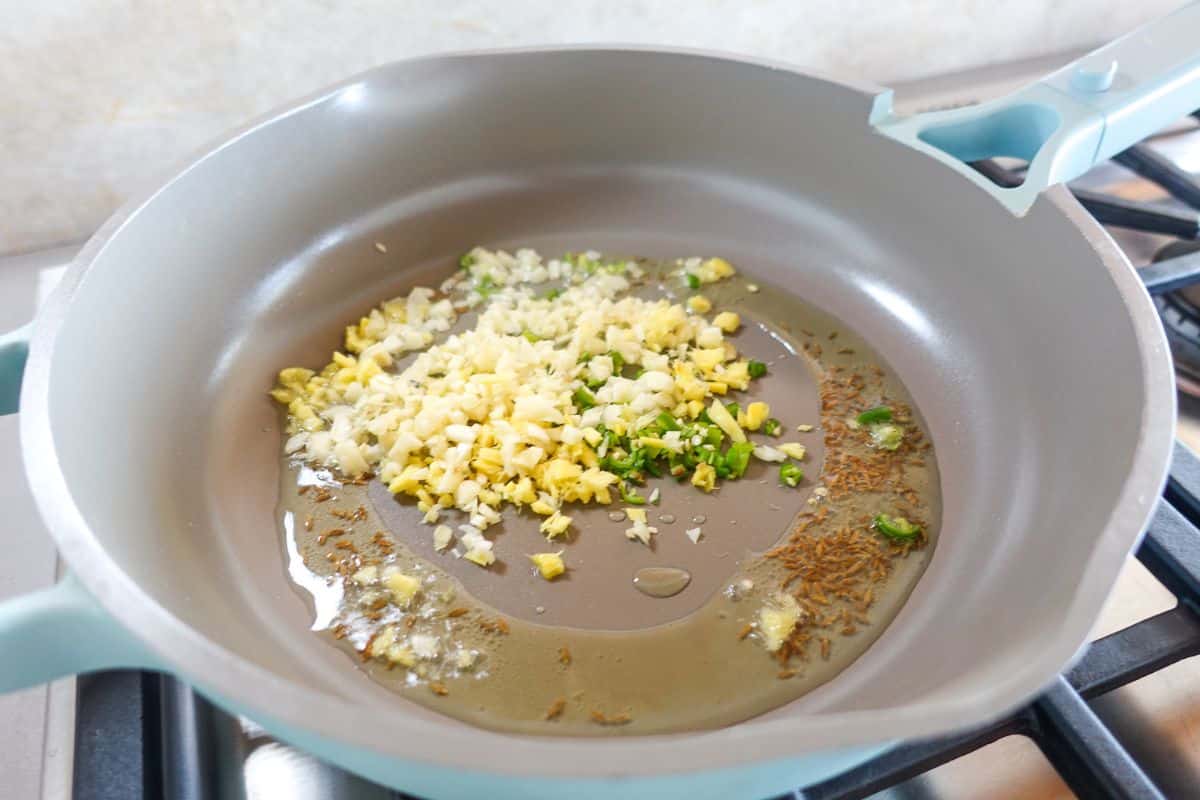 A frying pan on a stove contains oil, chopped garlic, ginger, green chili, and cumin seeds being sautéed.