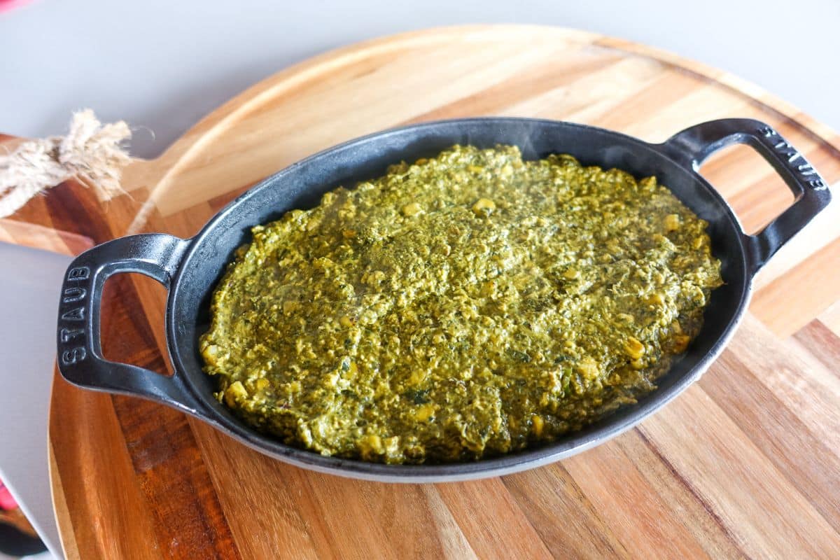 An oval black dish filled with palak paneer dip sits on a round wooden board.
