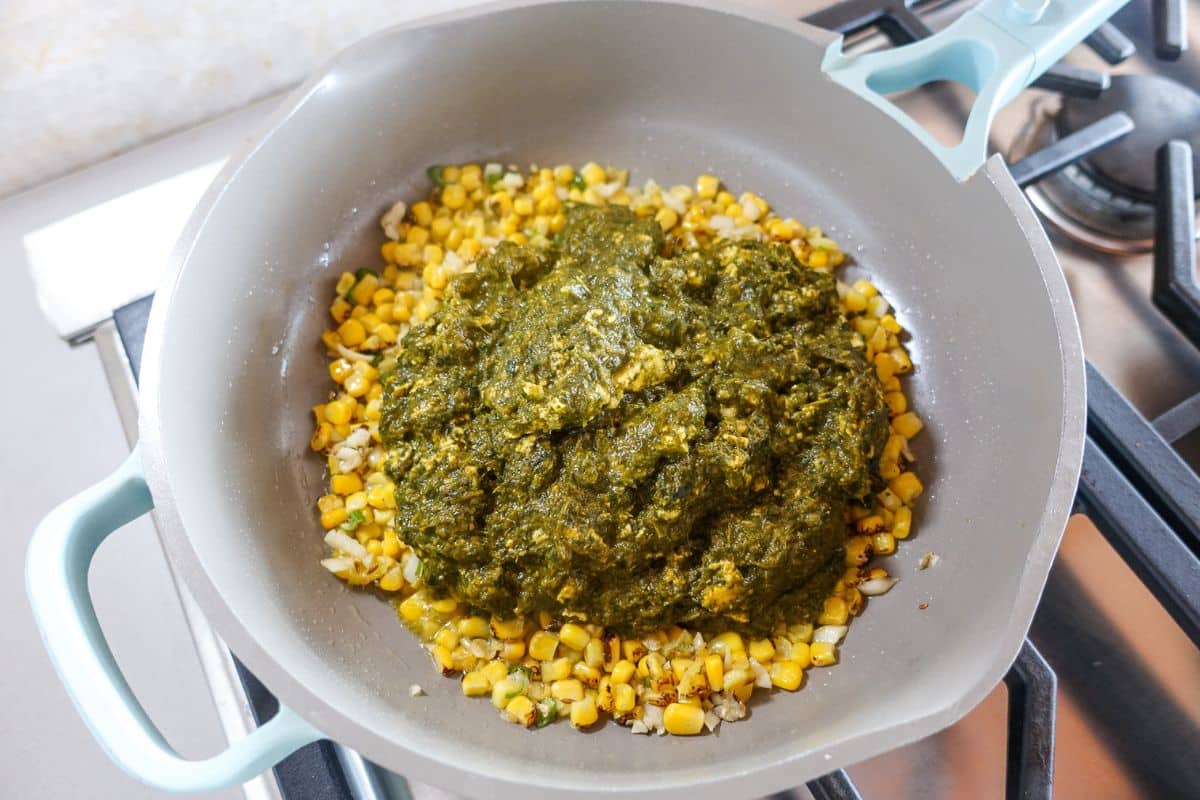A frying pan on a stove contains sautéed corn and palak paneer.