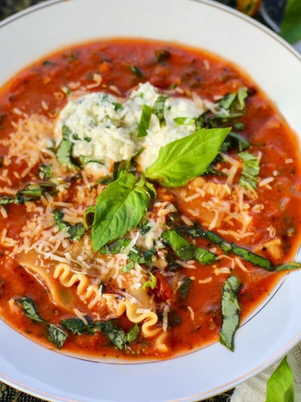 A bowl of vegetarian lasagna soup garnished with shredded cheese, fresh basil leaves, and two scoops of ricotta cheese, set on a decorative cloth with herbs scattered around.