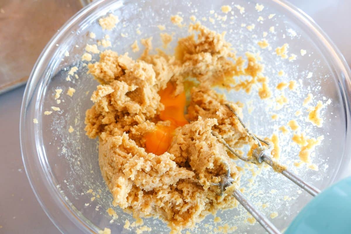 A glass bowl contains cookie dough with a cracked egg in the center and two metal beaters resting in the mixture.