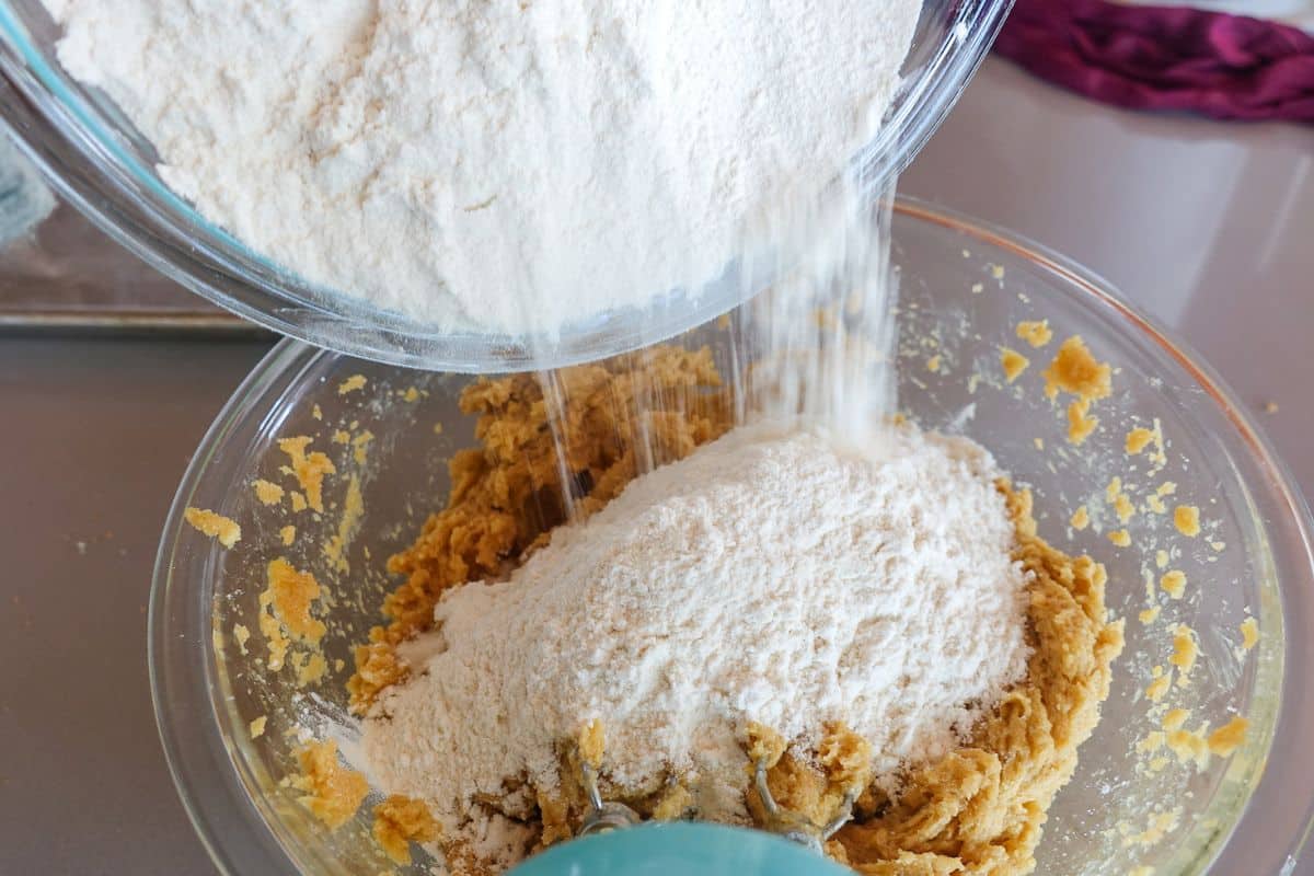 Flour is being poured from a glass bowl into a mixing bowl containing cookie dough on a kitchen counter.