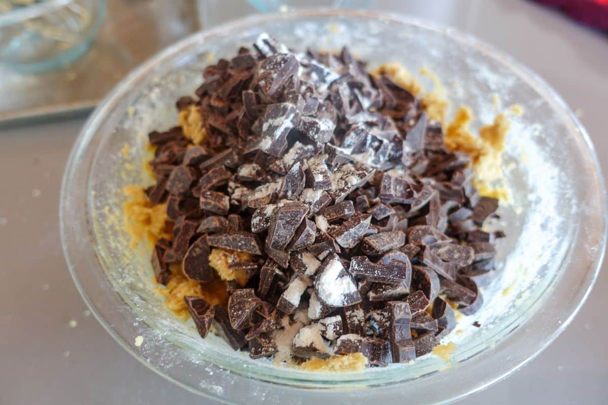 A glass bowl containing cookie dough topped with a large amount of chopped dark chocolate pieces and a dusting of flour.