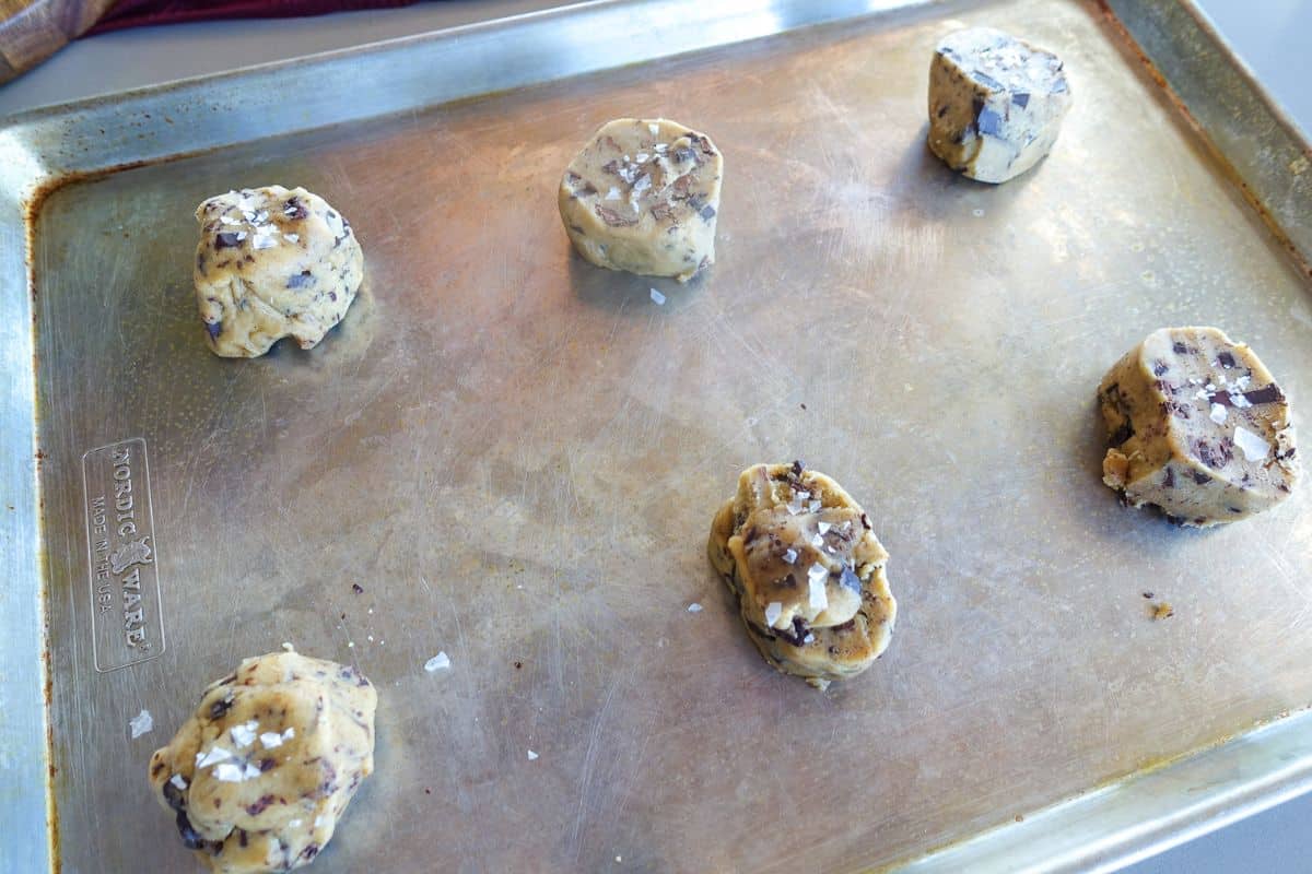 Six unbaked chocolate chip cookie dough balls, sprinkled with sea salt, are spaced out on a metal baking sheet.