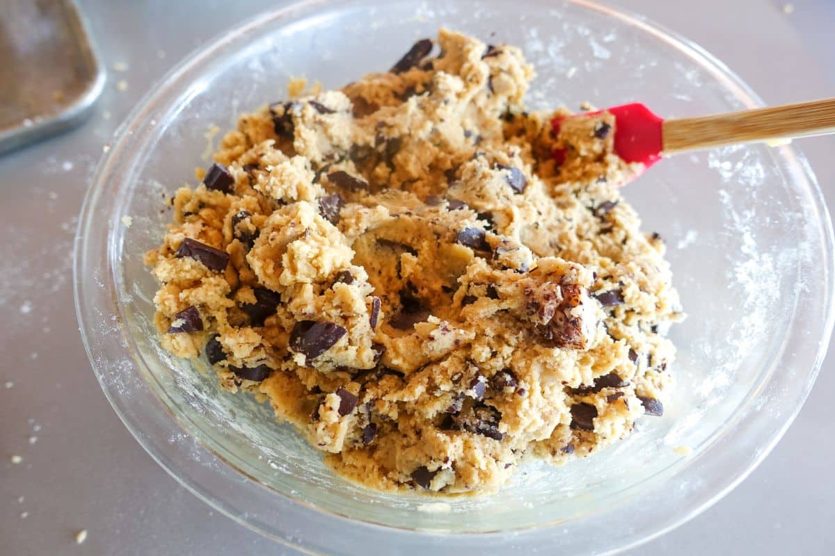 A glass bowl filled with chocolate chip cookie dough being mixed with a red spatula on a gray surface.