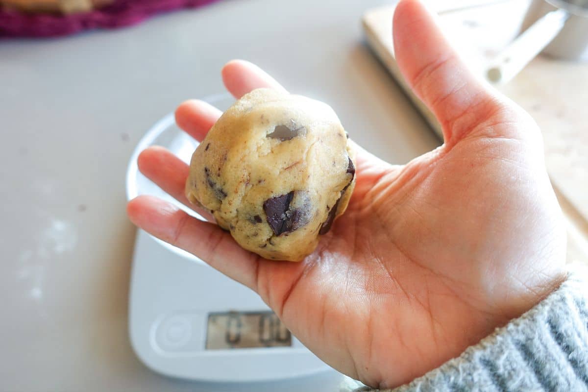 A hand holds a ball of chocolate chip cookie dough above a digital kitchen scale displaying numbers.
