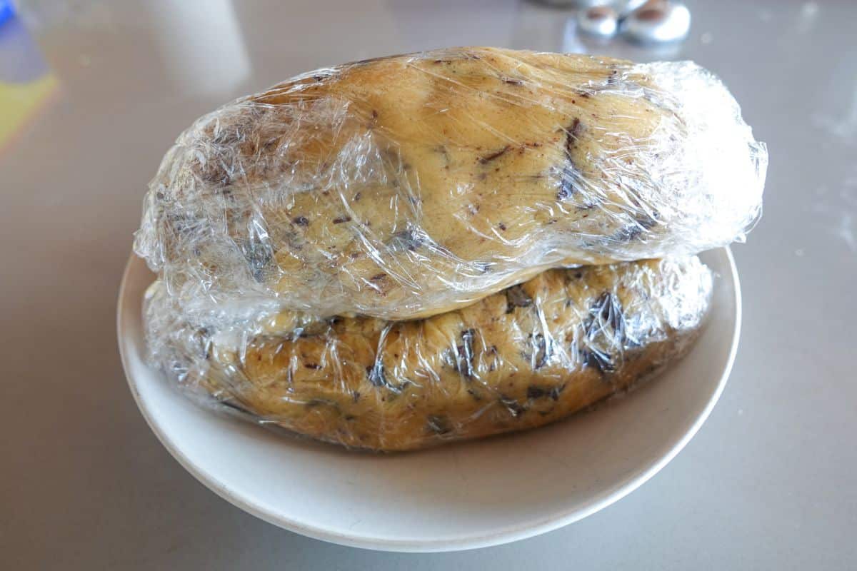 Two logs of cookie dough with chocolate chunks wrapped in plastic wrap are stacked in a white bowl on a countertop.