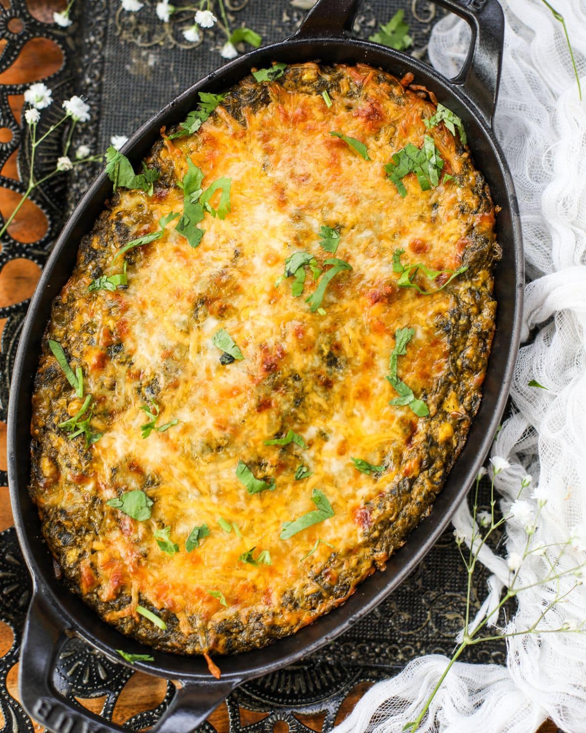 A baked palak paneer dip in a black oval dish, topped with melted cheese and chopped herbs, set on a patterned tablecloth with decorative flowers nearby.