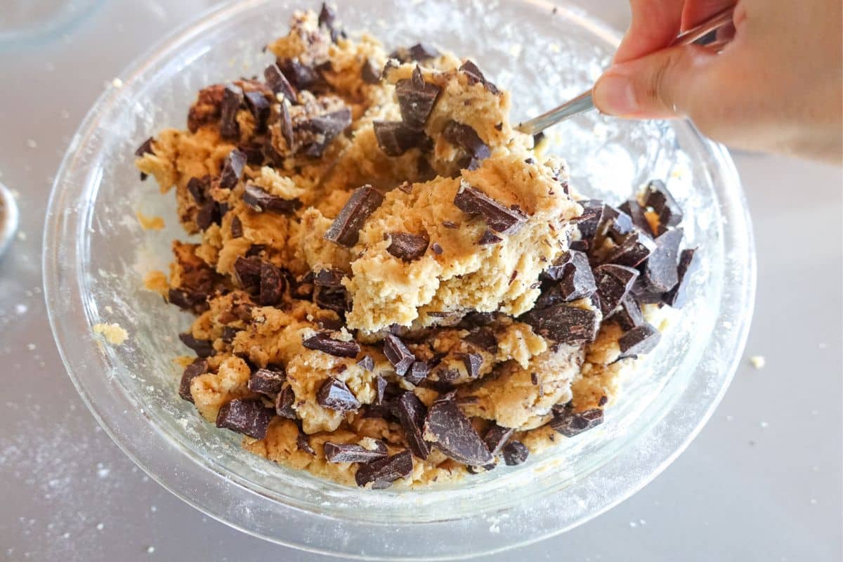 A hand uses a spoon to mix chocolate chunks into cookie dough in a clear glass bowl.