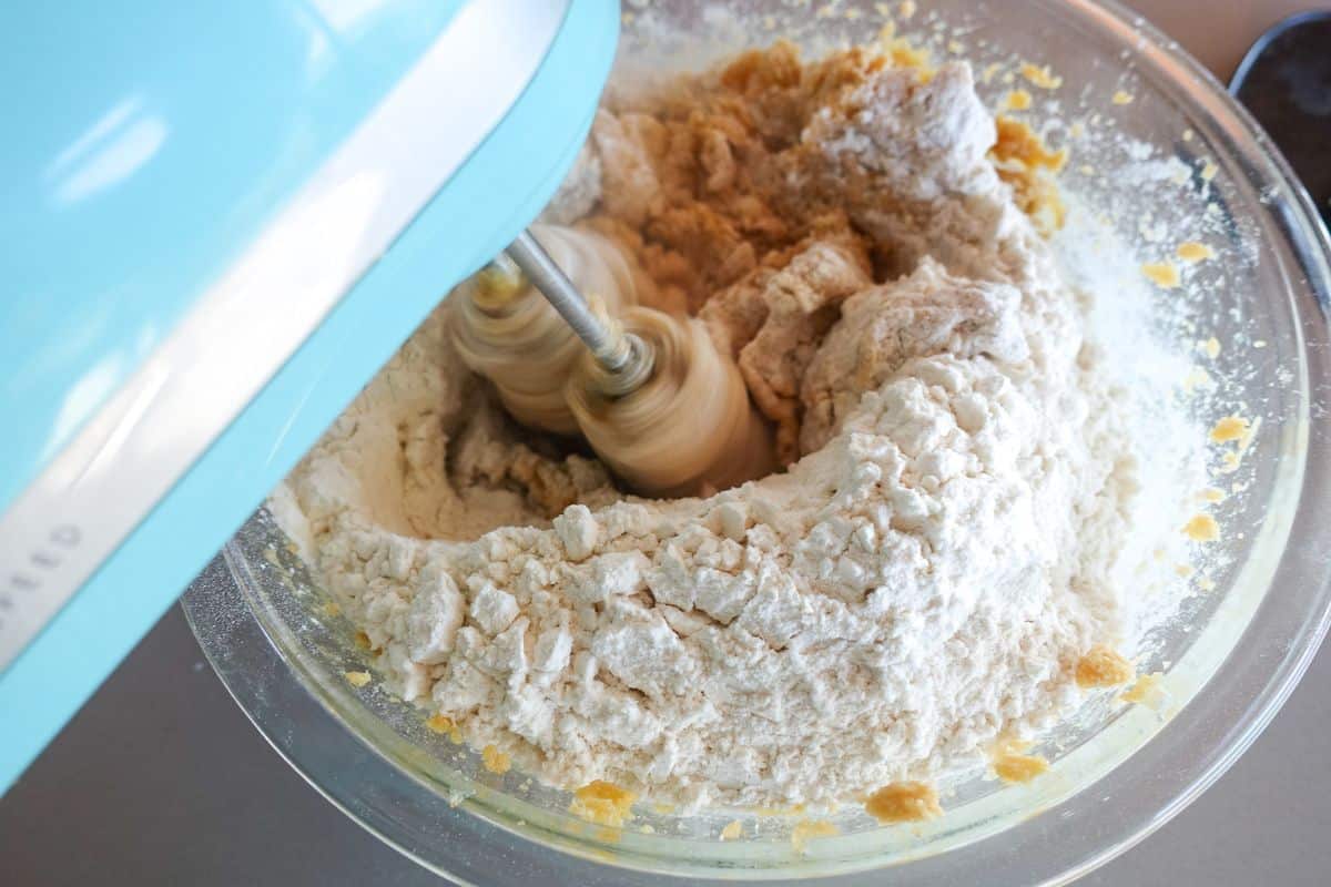 An electric mixer blends flour and other baking ingredients in a clear glass bowl.