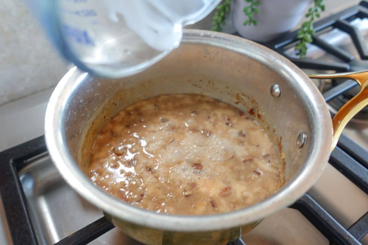 A saucepan containing a bubbling mixture is on a stove, with a measuring cup pouring almond milk into the pot.