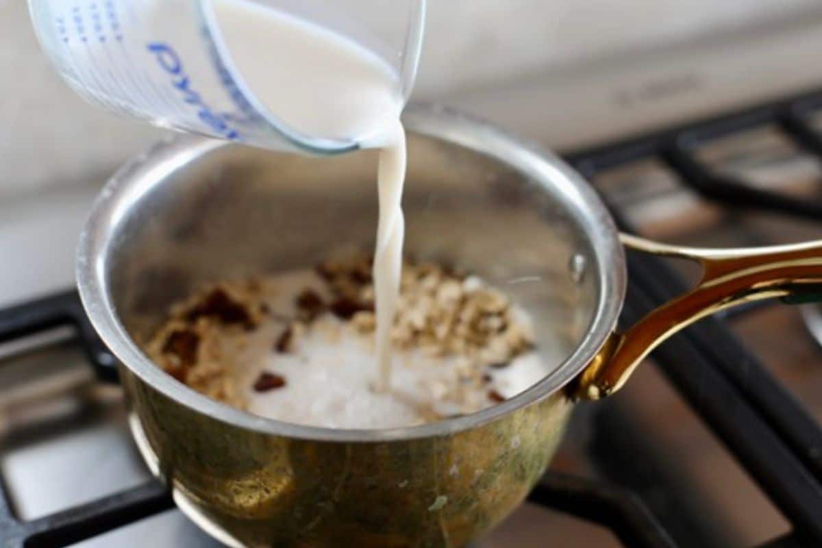 A measuring cup pours almond milk into a saucepan containing oats and other ingredients on a stovetop.