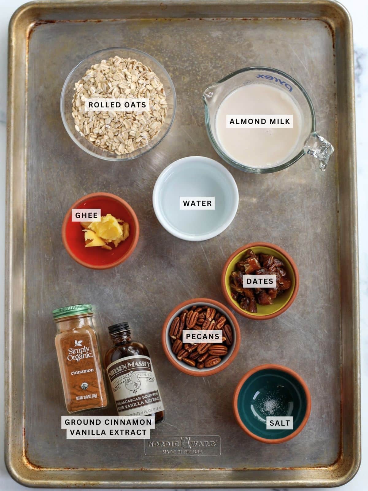 A baking tray with bowls containing rolled oats, almond milk, water, ghee, dates, pecans, salt, ground cinnamon, and vanilla extract, each labeled.