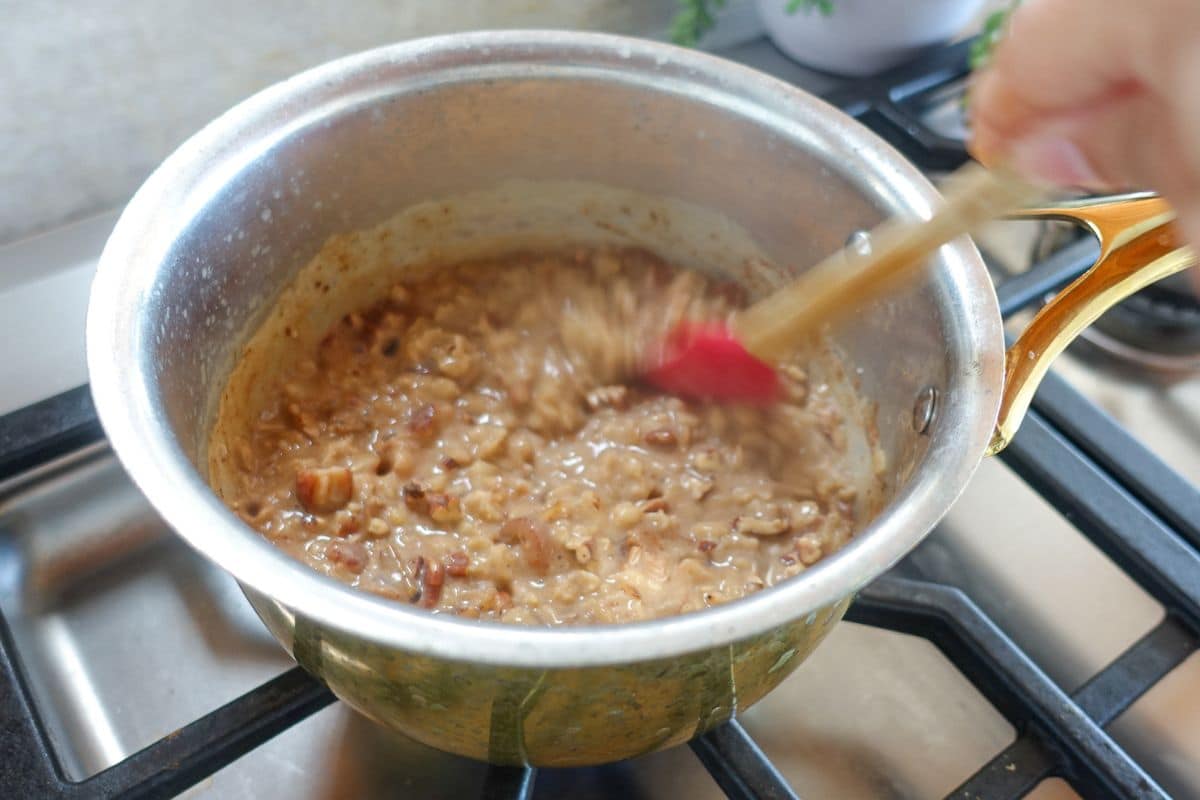 A hand stirs a pot of oatmeal with visible pieces of fruit or nuts, cooking on a gas stove.