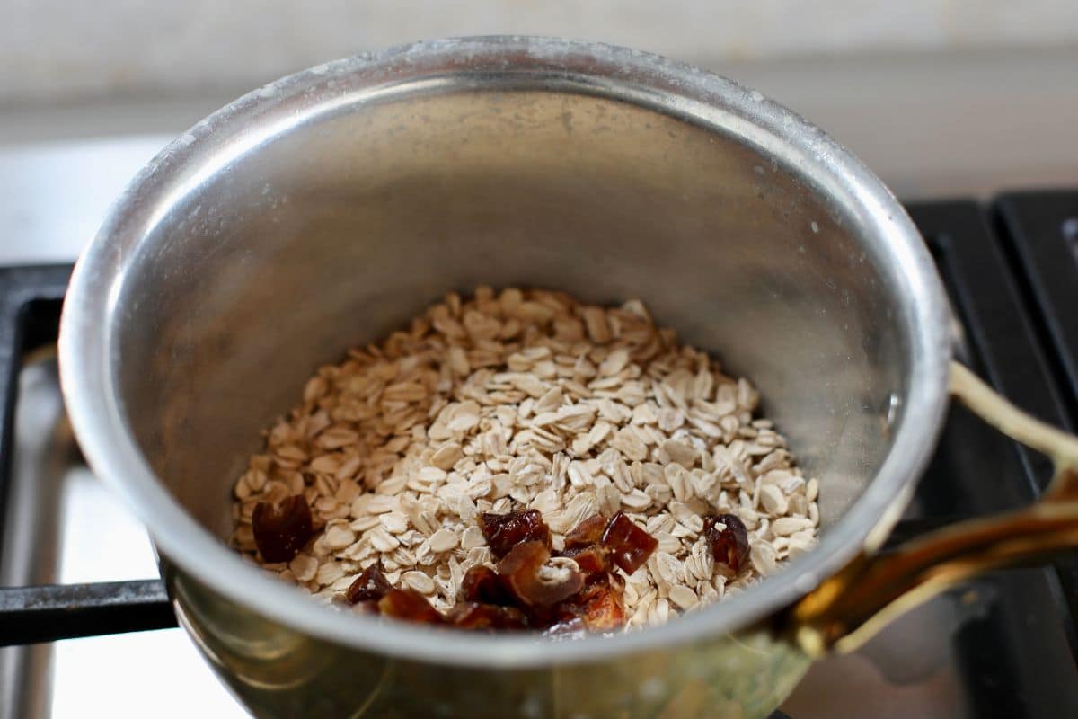 A metal saucepan on a stove contains dry rolled oats and chopped dates, ready to be cooked.