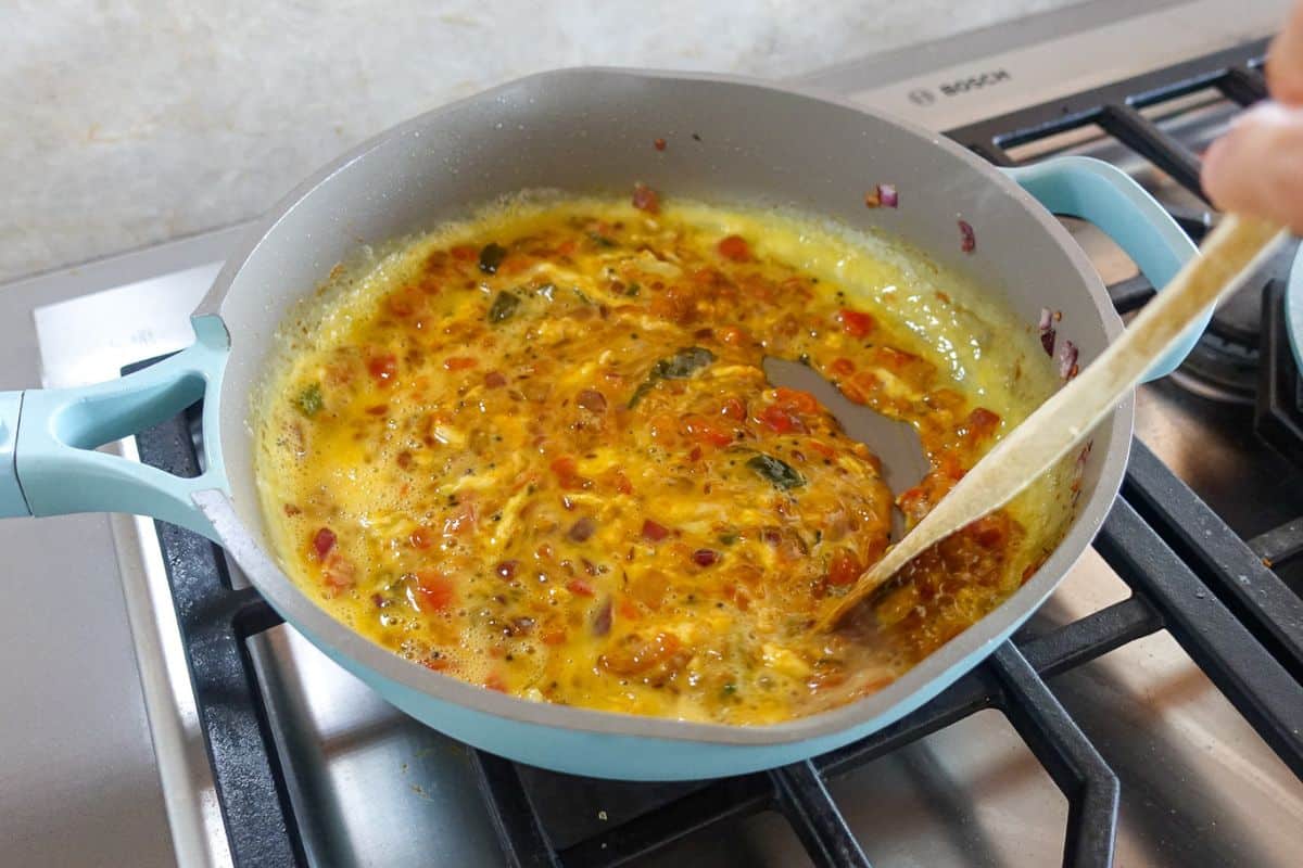 A wooden spoon is being used to scramble runny eggs in a pan.