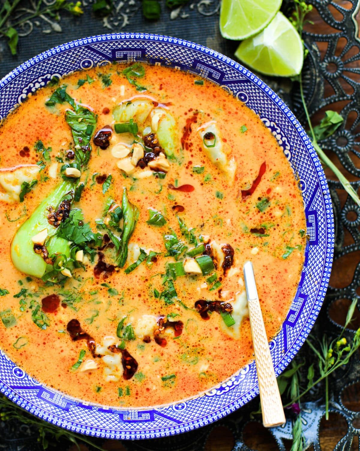 A bowl of spicy soup with bok choy, herbs, chopped nuts, and chili oil, served in a blue patterned dish with a spoon, and lime wedges on the side.