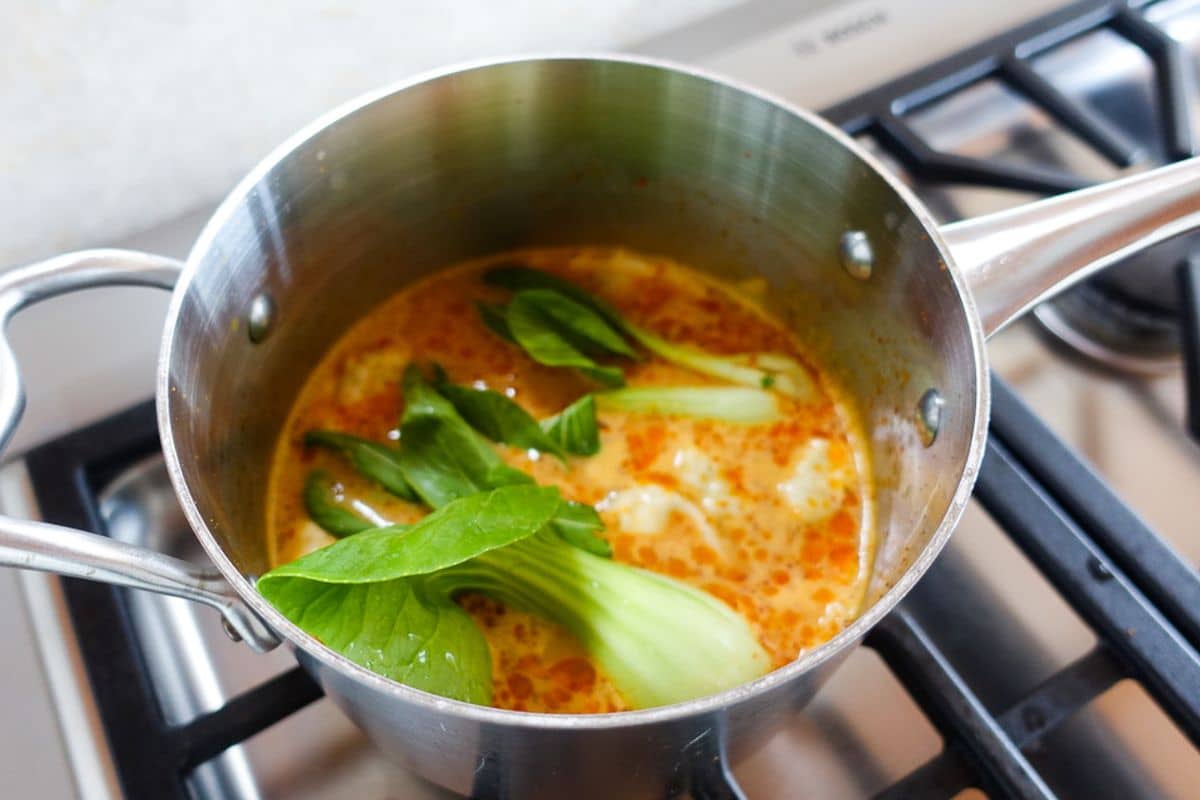 A stainless steel pot on a stove contains an orange broth with bok choy and other ingredients simmering inside.