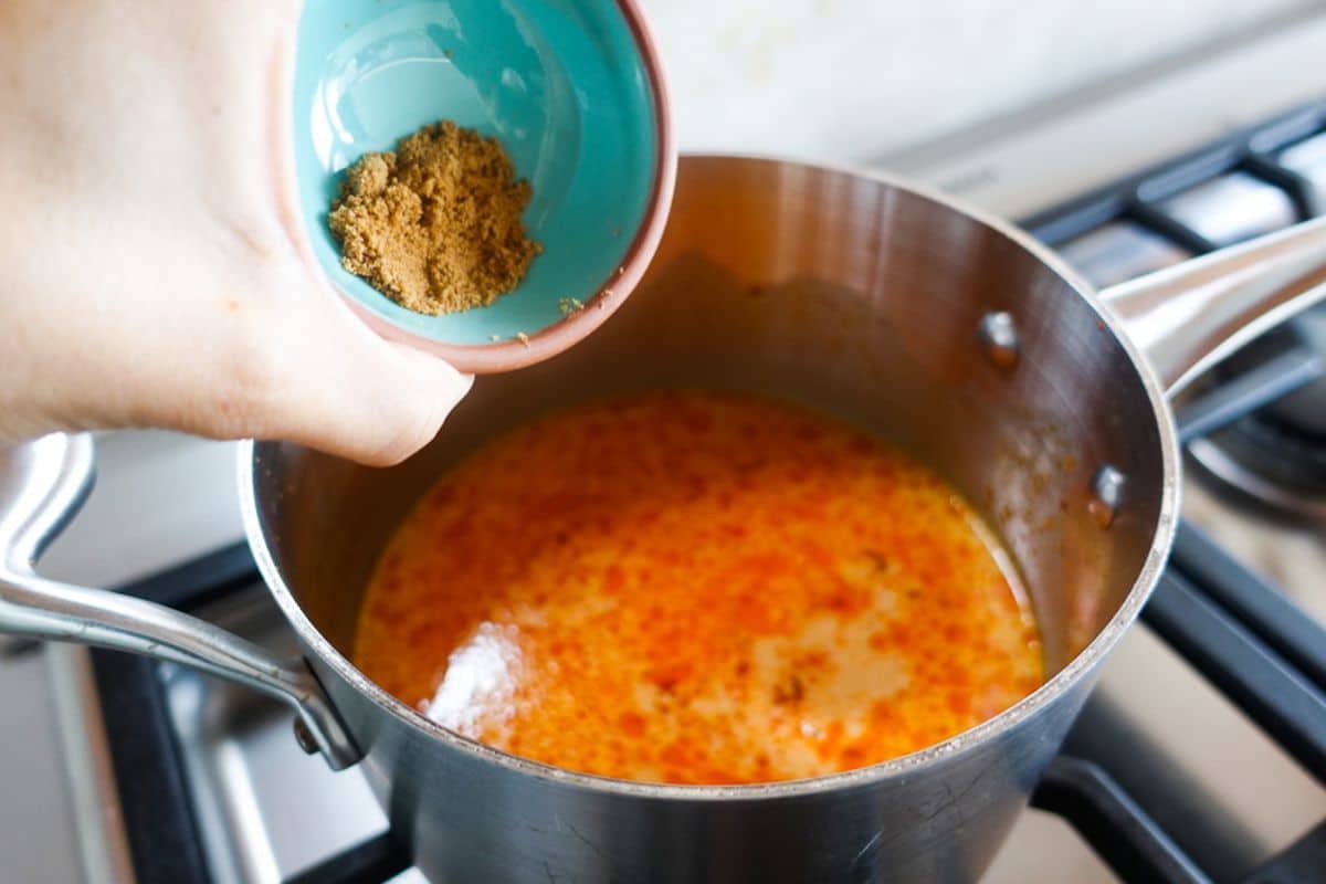 A hand holds a small bowl of coconut sugar above a pot of orange-colored liquid simmering on a stovetop.