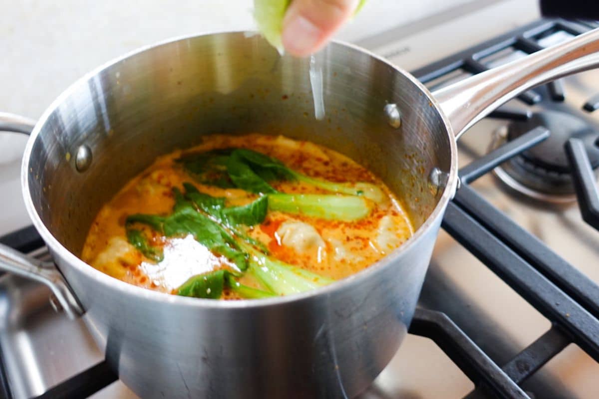 A hand squeezes lime over a pot of soup with greens and spices simmering on a gas stove.