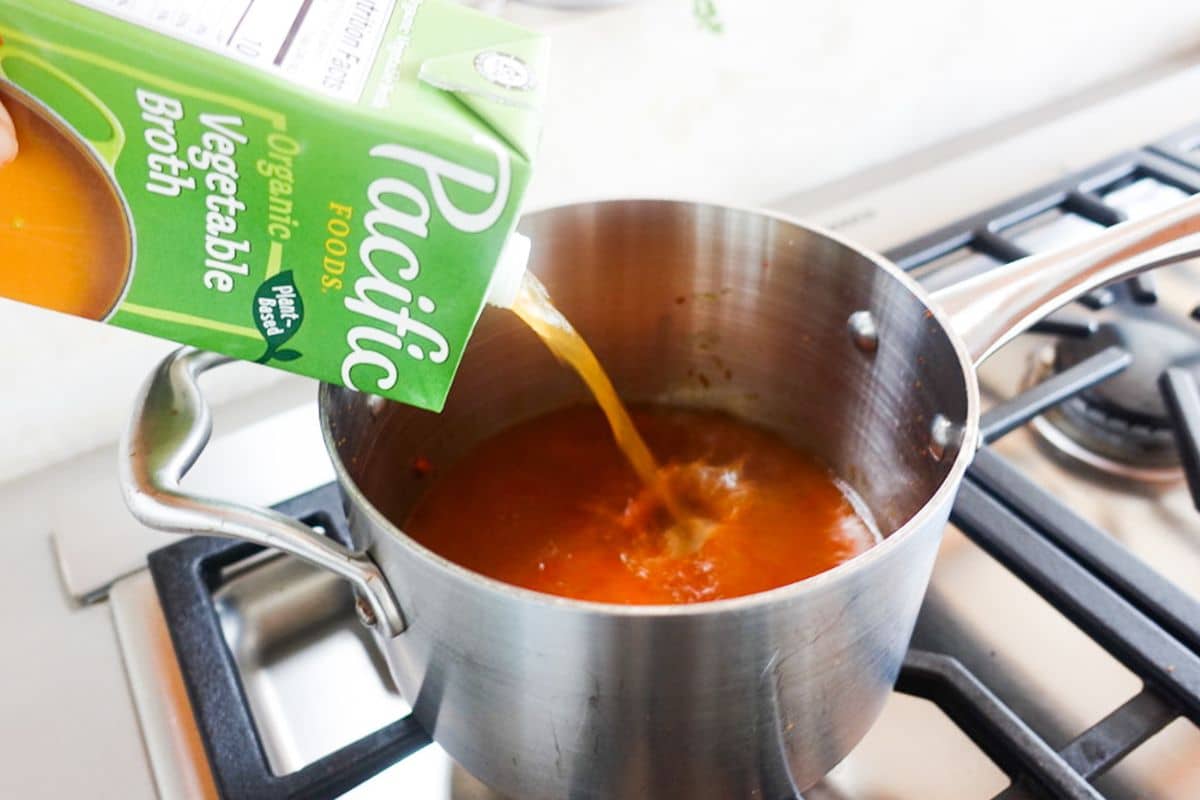A carton of organic vegetable broth is being poured into a pot with red soup on a stove.