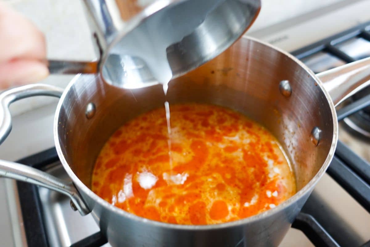 A hand pours coconut milk from a metal cup into a saucepan containing an orange-colored mixture on a stove.