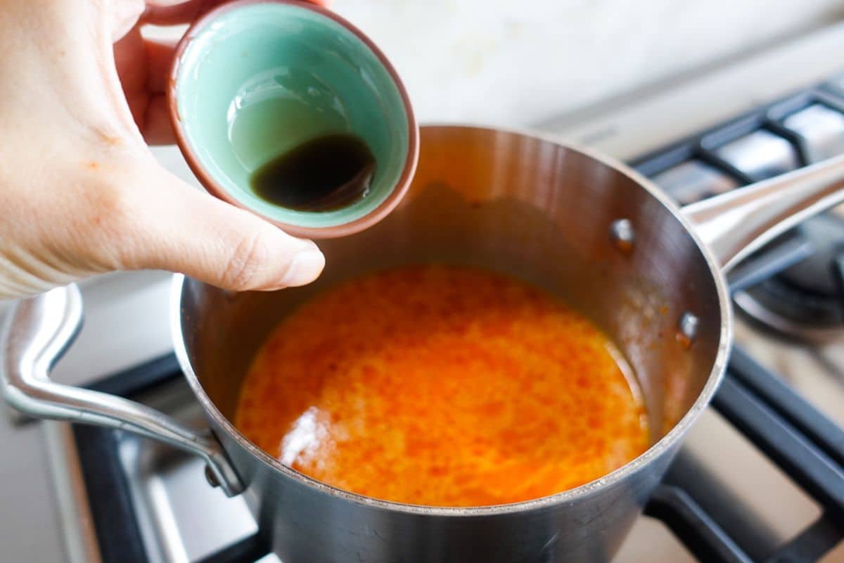 A hand holding a small bowl of fish sauce over a saucepan with orange-colored liquid simmering on a stove.