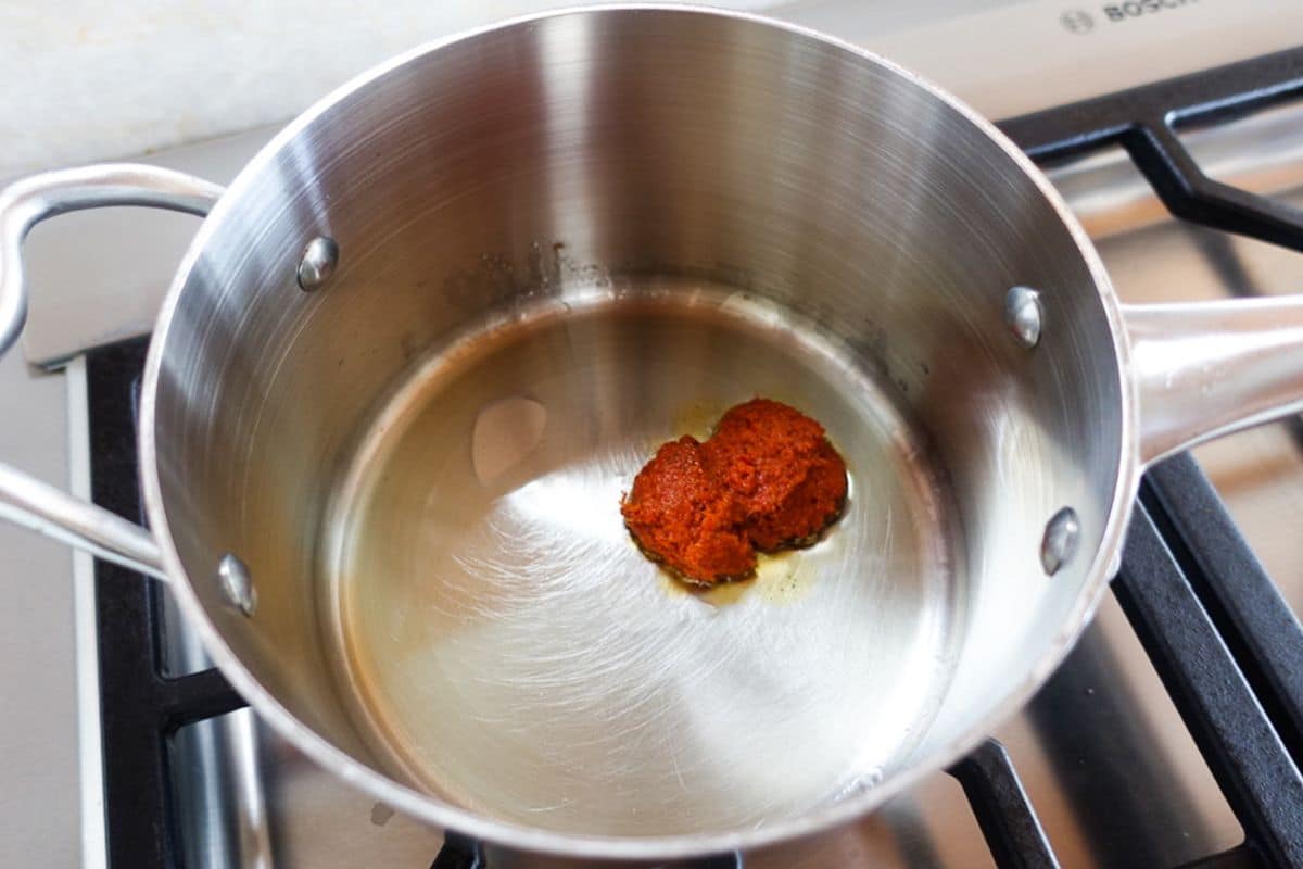 A stainless steel pot on a stove contains oil and a small amount of red curry paste.