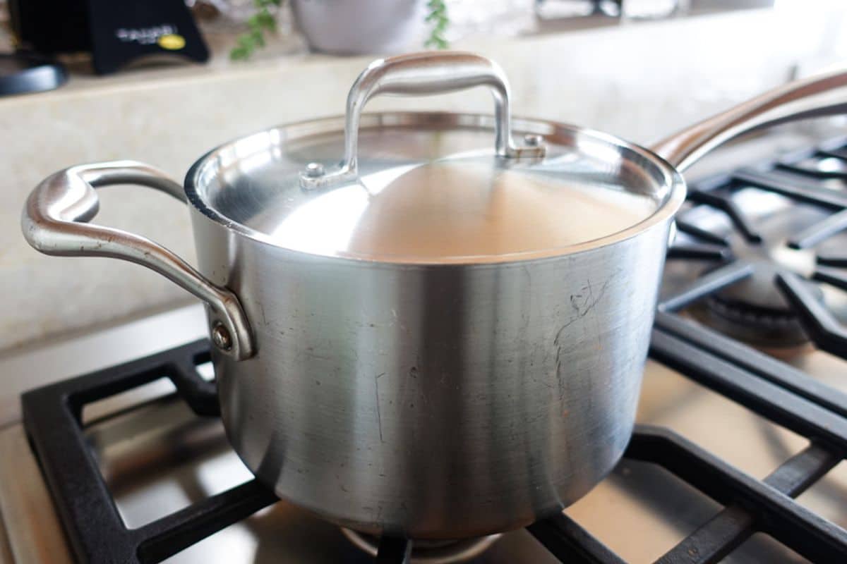 A stainless steel pot with a lid sits on a gas stove burner with the flame off.
