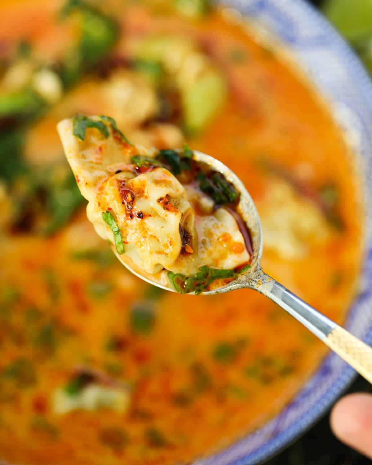 A close-up of a spoon holding a dumpling with herbs above a bowl of orange-colored soup.