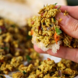 A hand holding a piece of bread topped with a spiced Indian scrambled eggs mixture containing cilantro, with more of the mixture and bread in the background.
