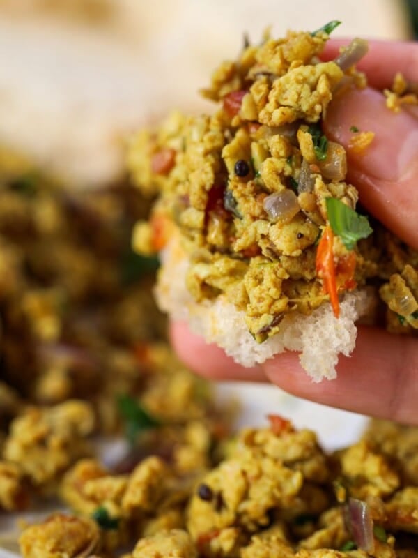 A hand holding a piece of bread topped with a spiced Indian scrambled eggs mixture containing cilantro, with more of the mixture and bread in the background.