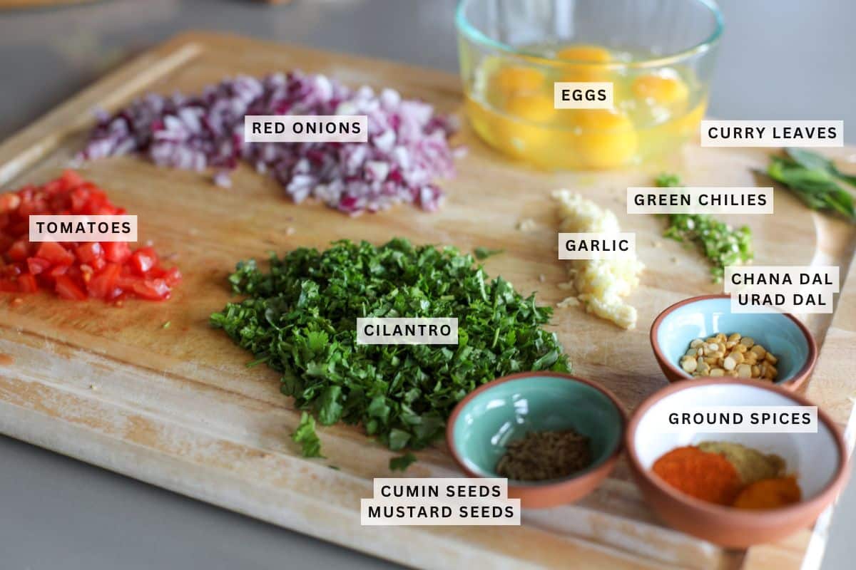 Chopped red onions, tomatoes, cilantro, garlic, green chilies, curry leaves, and various spices and dals are arranged on a wooden cutting board, with eggs in a glass bowl.